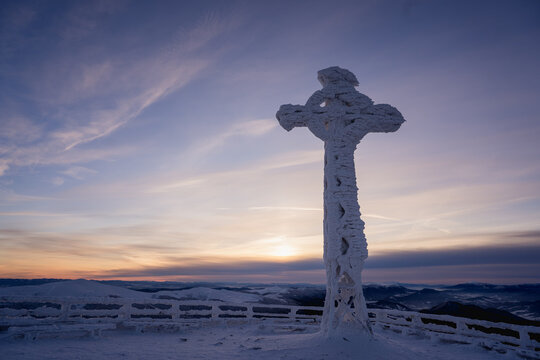 Frozen cross covered in rime ice on the summit of Tarnica in the Bieszczady Mountains during a cold winter sunrise. Snow-covered landscape, dramatic sky and peaceful mountain atmosphere.
