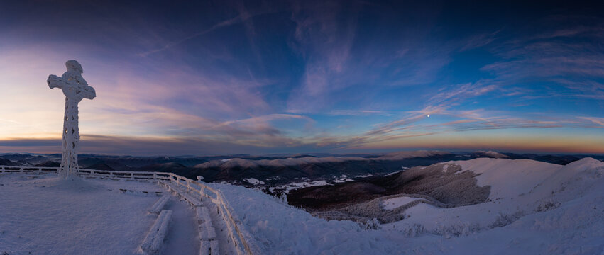 Frozen cross covered in rime ice on the summit of Tarnica in the Bieszczady Mountains during a cold winter sunrise. Snow-covered landscape, dramatic sky and peaceful mountain atmosphere.
 - Powered by Adobe