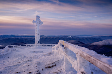 Frozen cross covered in rime ice on the summit of Tarnica in the Bieszczady Mountains during a cold winter sunrise. Snow-covered landscape, dramatic sky and peaceful mountain atmosphere.
