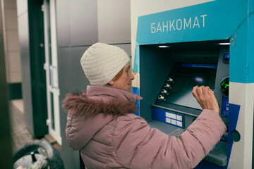 Senior woman using bank terminal withdrawing cash from ATM. Routine banking, interaction with technology, everyday financial independence and senior services.
