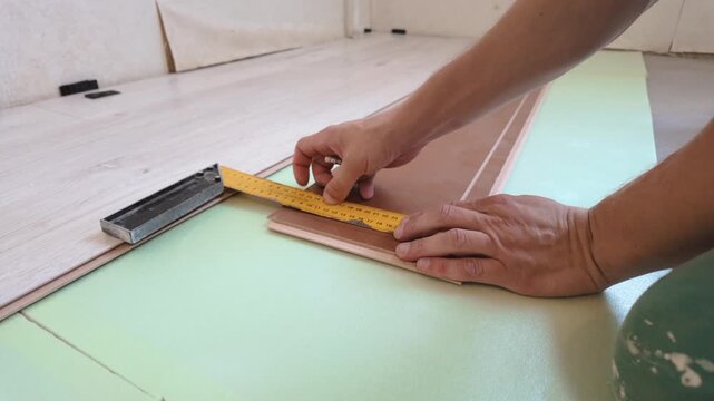 The craftsman's hands carefully measure the wooden laminate with a square ruler and pencil before cutting and installing new flooring during home renovation