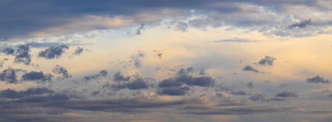 Panoramic view of evening sky with soft golden sun rays and scattered dark blue clouds during sunset