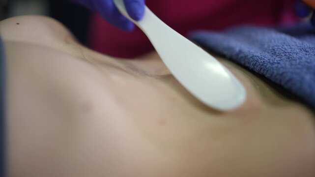 Caucasian woman chest waxing in spa, esthetician in gloves spreads warm wax with spatula across sternum over towel, closeup torso shot shows smooth skin, soft