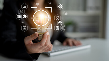A focused business professional holds a glowing light bulb symbolizing bright ideas. Digital icons surrounding the light bulb represent innovation and technology in a modern office. Gantry