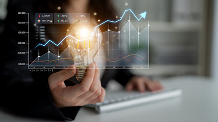 A businesswoman holds a light bulb symbolizing innovation while displaying digital graphs and charts that represent growth and data analysis in a modern office setting. Scalp