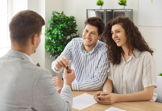 Smiling people receive key from realtor, excited couple and real estate agent sitting together at desk in office. Happy buyers sign home purchase contract, discuss agreement on meeting with broker