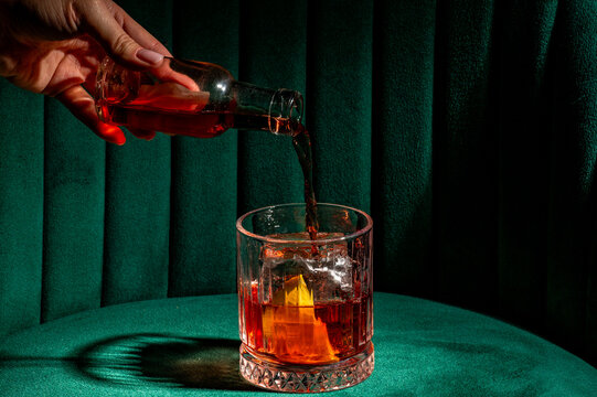 Person pouring a classic Negroni cocktail with gin, Campari, and sweet vermouth into a rocks glass with ice and an orange peel on a vibrant green velvet background