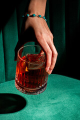A close-up shot of a hand with a turquoise bracelet reaching for a vibrant red cocktail in a rocks glass, against a plush green velvet background.