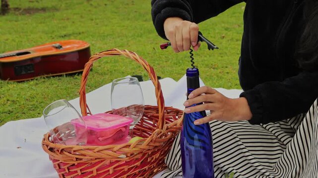 Woman using a corkscrew to open a bottle of wine during a picnic, representing romance, leisure, celebration, and shared moments in an outdoor natural setting.