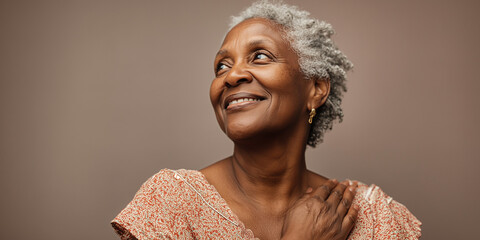 Portrait of a happy senior African American woman with grey hair smiling and looking away while holding her hand to her chest against a brown background.