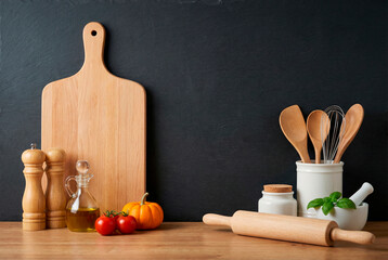 Kitchen countertop with cutting board, utensils, tomatoes, pumpkin, oil, and spices on a wooden table against a dark background.