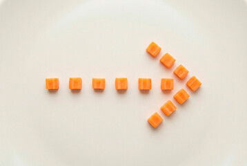 A creative arrangement of chopped carrot cubes forming an arrow on a clean white surface for healthy eating and food presentation concepts.