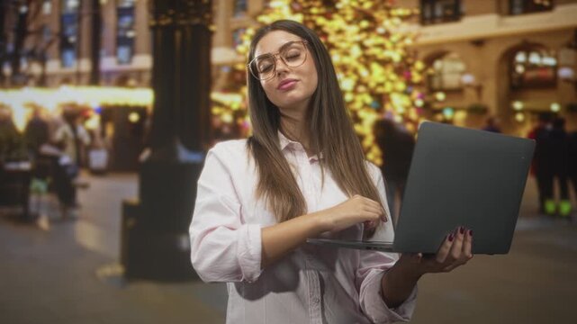 Woman with glasses holds laptop, types with right hand and tucks hair while standing by a lit christmas tree and blurred crowd in a building; confidence focus.