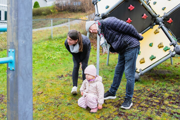 Family members, a woman and a man, are observing a young child playing on a playground structure, surrounded by grass and a fence, capturing a joyful moment in outdoor leisure