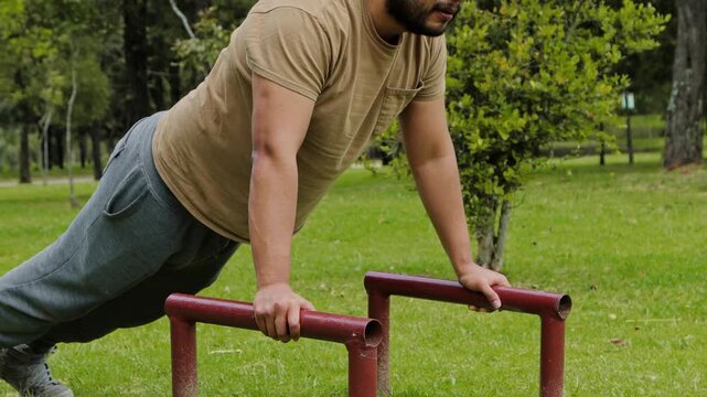 Man performing push ups on low parallel bars placed on the ground, showing upper body strength, effort and bodyweight training in an outdoor workout.