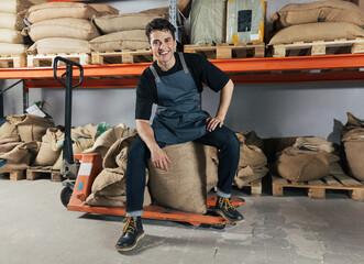 Confident and cheerful warehouse worker sitting on bag. Young smiling male worker in an apron...