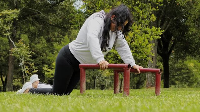 Woman attempting to perform push ups on low parallel bars, showing effort, difficulty and determination during an outdoor bodyweight workout.