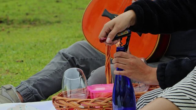 Young couple at a camping site where the woman opens a cork wine bottle using a corkscrew while the man holds a guitar, representing leisure, music and outdoor lifestyle.