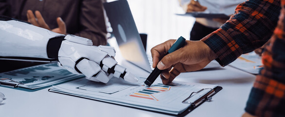 A futuristic scene showing a human and a robotic hand engaged in a collaborative meeting while analyzing data charts and visual presentations in a modern office setting.Noogenesis