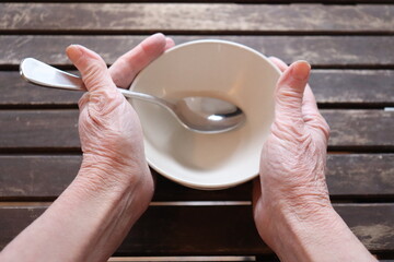 A old woman holding an empty bowl on a old wood dining table. Old hands holding an empty dish to eat. The price of food and the poverty among older adults. Cost of the living and elders.