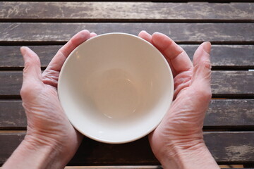 A old woman holding an empty bowl on a old wood dining table. Old hands holding an empty dish to eat. The price of food and the poverty among older adults. Cost of the living and elders.