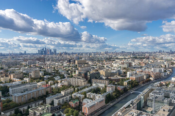 Obraz premium A breathtaking aerial view captures a sprawling Moscow cityscape filled with diverse buildings, green spaces, and a winding river. Fluffy clouds drift across the blue sky, enhancing the view.