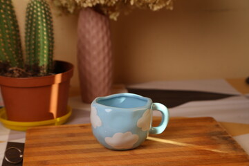 Cute ceramic cup with cloud design placed on wooden cutting board near potted plant in bright kitchen