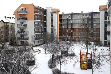snowy winter scene of modern condominium building. court yard with bare trees and playground. swings and slide. exterior corridors. metal picket railing. stucco elevation with windows. home concept