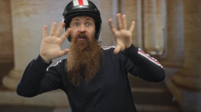 Man wearing helmet and track jacket raises both hands calling out vocally in front of building columns; alertness.