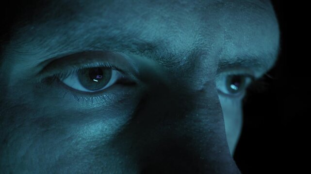 Male eyes stare intensely at a computer screen with digital reflections in the pupils. Cool blue lighting illuminates the face in a dark environment suggestive of hacking or gaming.