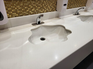 An unusual shaped white sink in a public toilet. Chrome-plated high-pressure washbasin faucets.