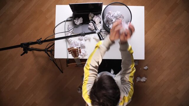 Male songwriter discarding numerous crumpled papers into a trash can, experiencing creative block while working on new music with his laptop, microphone, and guitar
