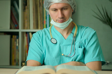 A doctor or medical worker in uniform with face mask reads a book at his desk. Stethoscope on neck. Concept of medical education, professional development and student training.