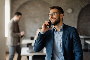 Professional businessman talking on a smartphone in a modern open-plan office, with a diverse team of colleagues working in the background.
