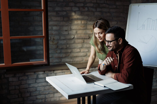 Two young creative professionals collaborating on a project while using a laptop in a modern design studio, sharing ideas and working together in a bright office space - Powered by Adobe