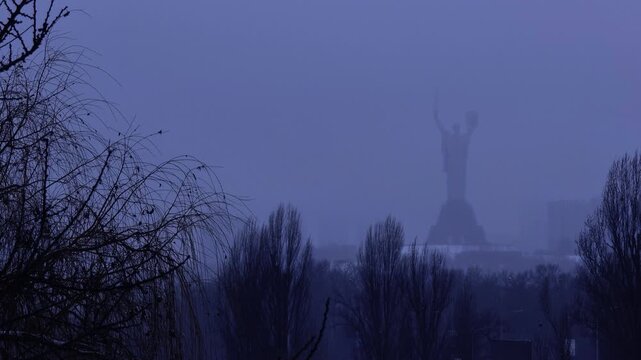 Iconic Mother Ukraine monument in Kyiv standing tall amidst a thick winter fog at dusk, with snow-covered trees in the foreground, creating a mysterious and atmospheric cityscape scene