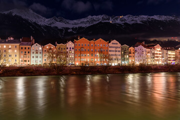 Traditional colourful residential buildings on the bank of a river overlooked by towering snow-capped mountains at night in winter. Innsbruck, Austria,