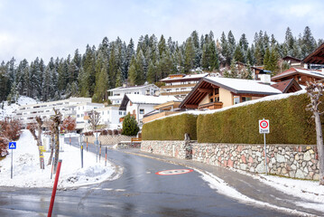 Holiday apartments and houses covered in snow along a curvy street in a mountain village in the Alps in winter. A snowy forest is visible in background.