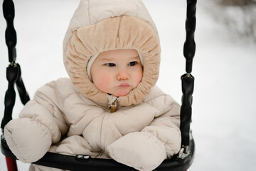 A curious baby, bundled in a snug snowsuit, experiences winter fun on a swing. The scene captures the delight of a child or toddler exploring the snowy outdoors with wonder and joy.