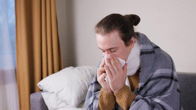 Sick man wrapped in a blanket blowing his nose and using nasal spray. Male with cold or flu symptoms treating nasal congestion and rhinitis at home. High quality 4k footage