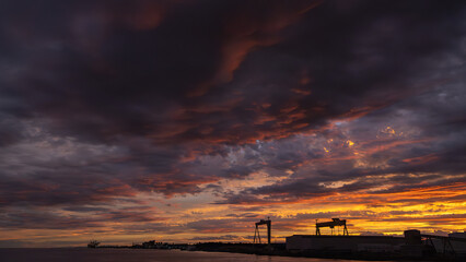 Industrial harbor silhouette at sunset with a dramatic, fiery orange and purple sky. Cargo cranes and port infrastructure under a moody, textured cloudscape