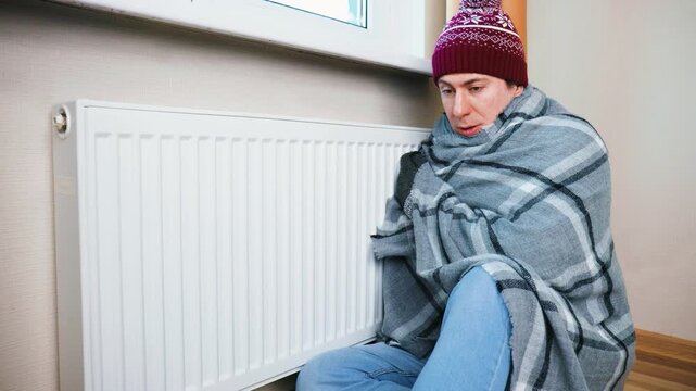 Freezing man in winter hat and blanket shivering near a radiator. Concept of heating problems, energy crisis, rising gas prices, and cold temperature in the apartment. High quality 4k footage