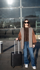 Obraz premium Young woman wearing sunglasses, a brown sweater, plaid coat, and jeans, walking with a black suitcase along the pavement in front of a modern glass building, representing travel and departure