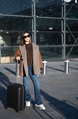 Woman standing outdoors near a glass facade, wearing sunglasses and warm stylish clothes, holding a black suitcase, ready for a journey or urban exploration