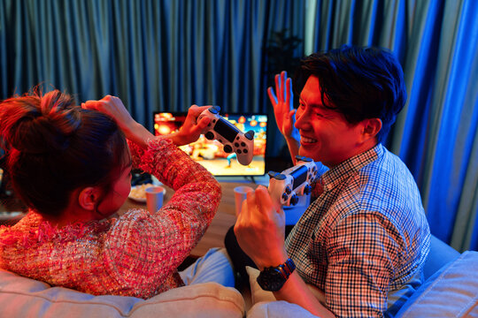 Couple gamer with hands cross of joystick playing fighting video game with winner together on tv screen at back side view, sitting sofa at neon light color living room at modern comfy home. Infobahn.