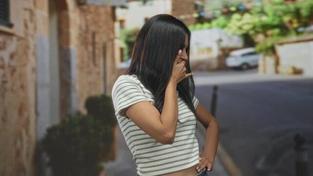 Woman covers mouth with hand on narrow street, hand on hip, striped tshirt and jeans visible; playful mischief.