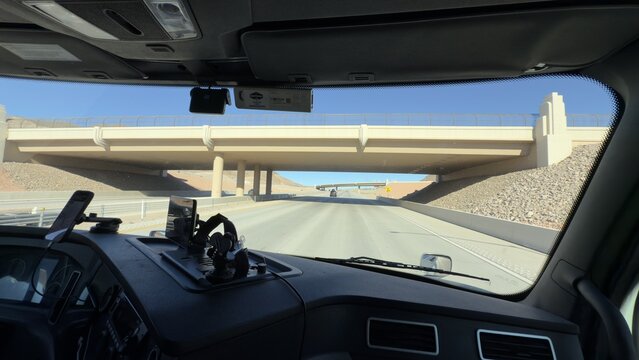  View from a truck cabin driving on a highway under two sunlit bridges on gravel embankments. Visible dashboard inside. Bright sunny day atmosphere of a professional road trip.