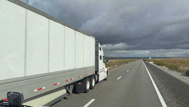 White Semi-Truck Overtaking on Highway Viewed from Another Truck Cabin POV. Overcast sky with grey clouds, distant road stretching to the horizon, and dry winter fields in Nevada or Utah.
