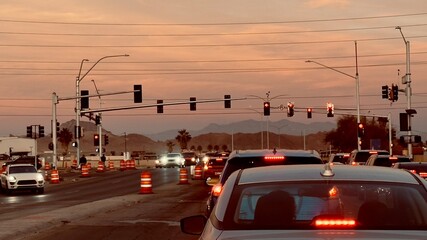POV from inside a car at a city intersection during golden hour. Vehicles with glowing tail lights wait at a red light against a backdrop of glowing orange mountains and clouds in Nevada.