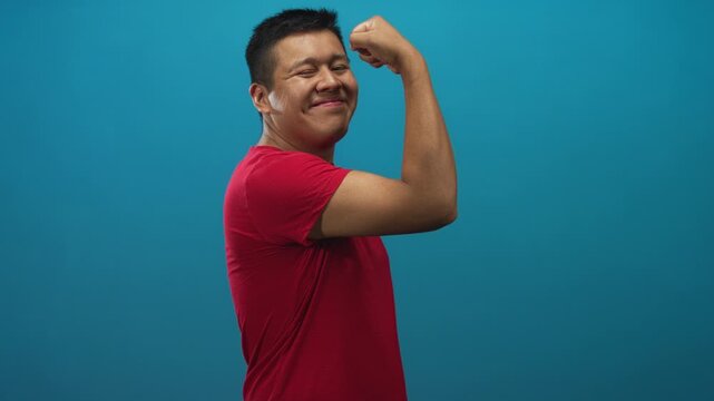 Hispanic man flexes bicep and smiles wearing red t shirt, side pose in studio with teal backdrop; confidence strength.